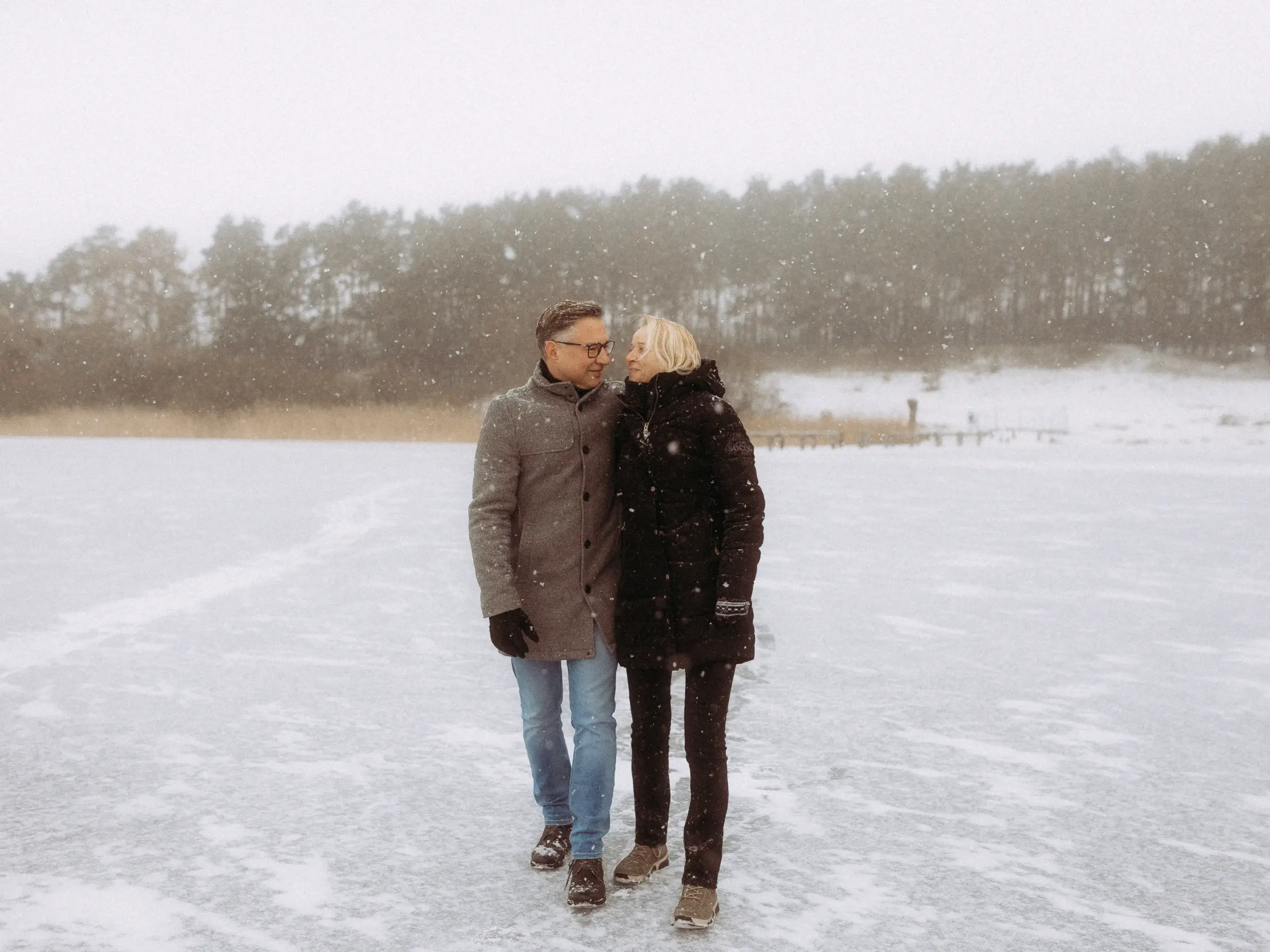 Ein Paar spaziert eng umschlungen über einen zugefrorenen, verschneiten See bei leichtem Schneefall, fotografiert bei einem Paarshooting im Winter von Jan Büchler.