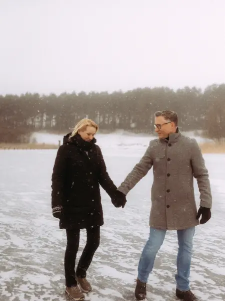 Ein Paar hält händchenhaltend inne bei einem romantischen Winterspaziergang auf einem zugefrorenen See, während es sanft schneit. Die beiden blicken sich lächelnd an, umgeben von einer verschneiten Waldkulisse. Authentische Pärchenfotos Schwerin in winterlicher Atmosphäre.