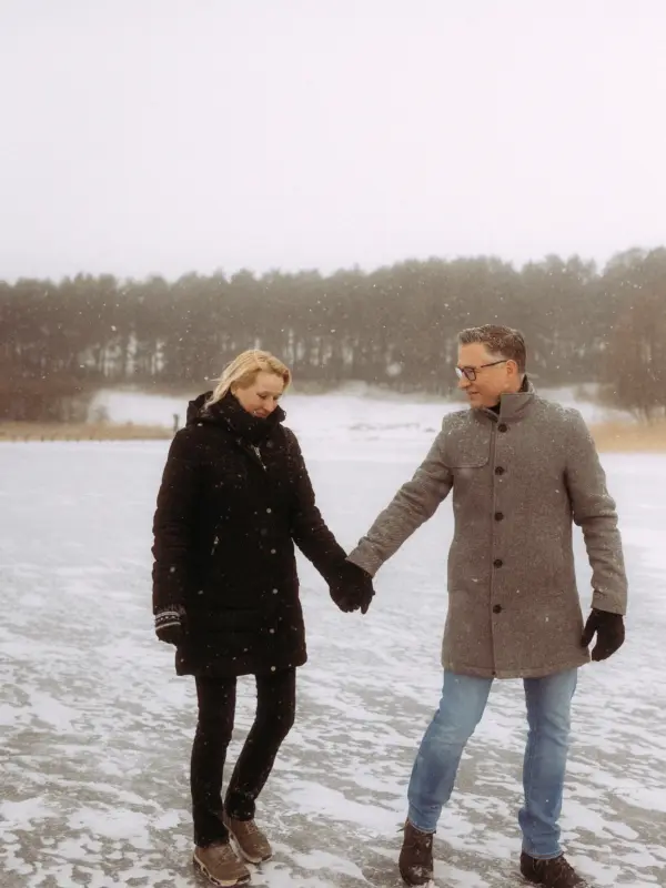 Ein Paar hält händchenhaltend inne bei einem romantischen Winterspaziergang auf einem zugefrorenen See, während es sanft schneit. Die beiden blicken sich lächelnd an, umgeben von einer verschneiten Waldkulisse. Authentische Pärchenfotos Schwerin in winterlicher Atmosphäre.