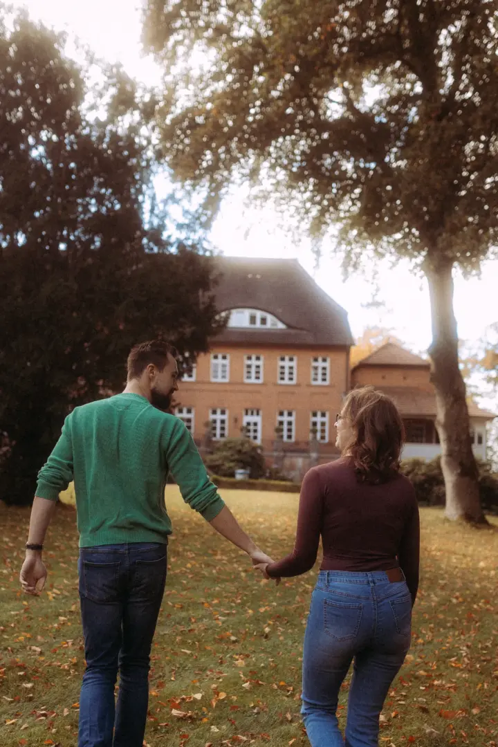 Ein Paar spaziert Hand in Hand durch einen herbstlichen Park vor einem historischen Herrenhaus, festgehalten von einem Fotograf für Pärchen in Schwerin. Die beiden blicken sich lächelnd an, während das warme Sonnenlicht durch die Bäume fällt und eine romantische Stimmung erzeugt.
