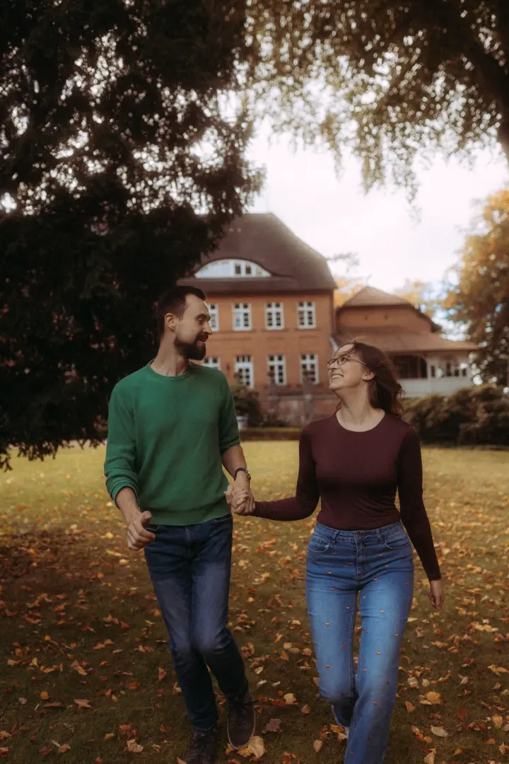Ein glückliches Paar bei einem romantischen Spaziergang in einem herbstlichen Park vor einem historischen Backsteingebäude. Die beiden halten Händchen und lachen sich an, während sie über das Laub laufen. Professionelle Pärchenfotos Schwerin in natürlicher, stimmungsvoller Atmosphäre.