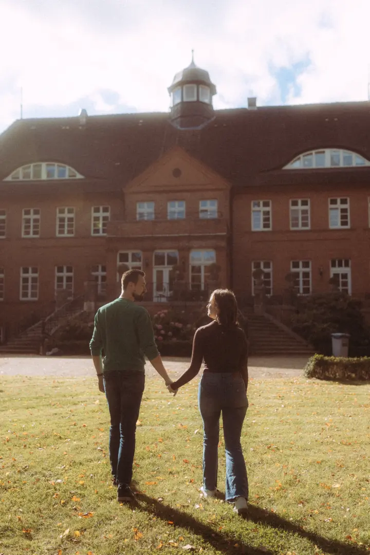 Ein Paar hält Händchen und geht gemeinsam über eine sonnige Wiese vor einem historischen Backsteingebäude. Diese romantischen Pärchenfotos in Schwerin fangen einen harmonischen Moment in einer herbstlichen Kulisse ein, ideal für eine Lovestory als Fotograf in Schwerin und MV.