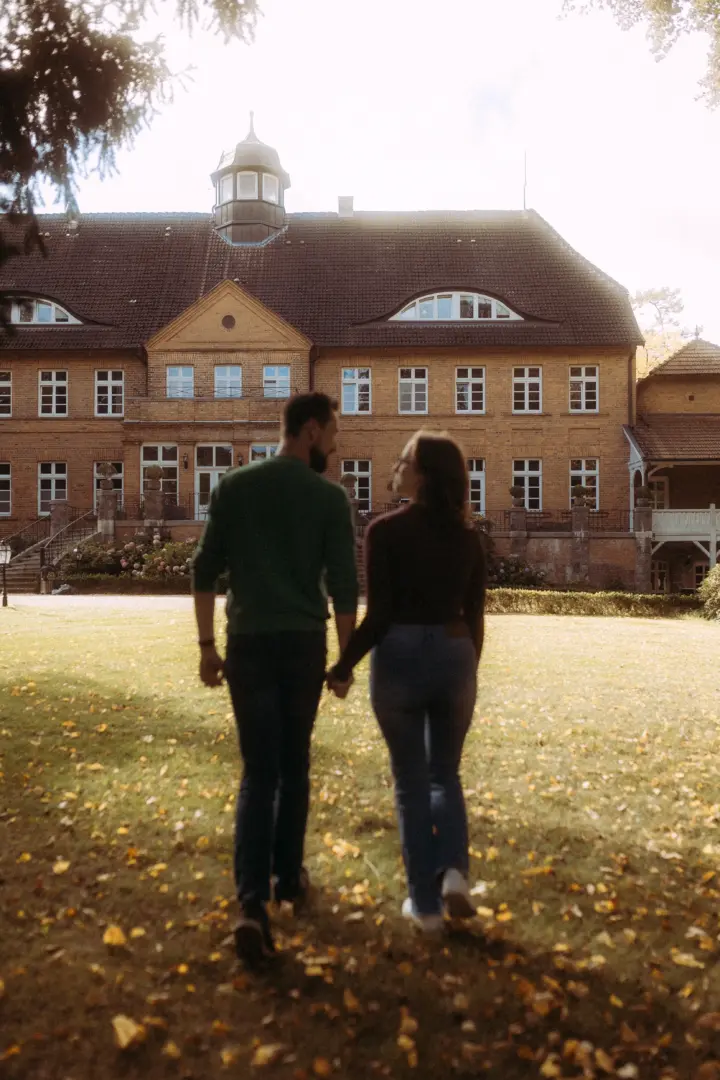 Ein Paar spaziert Hand in Hand über einen herbstlichen Rasen vor einem historischen Herrenhaus. Die romantische Stimmung unterstreicht die professionelle Arbeit als Fotograf für Pärchen in Schwerin, während das Paar in herbstlicher Kleidung auf das imposante Gebäude zuläuft.