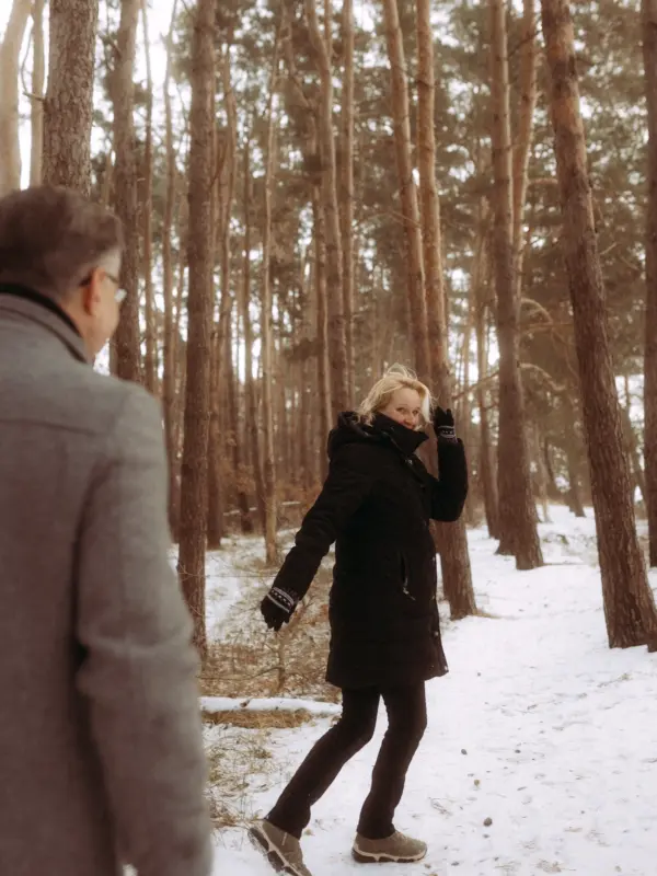 Eine blonde Frau in schwarzer Winterjacke blickt lächelnd über die Schulter zu ihrem Partner, während sie gemeinsam durch einen verschneiten Kiefernwald spazieren. Diese authentischen Pärchenfotos in Schwerin fangen einen romantischen Wintermoment in der Natur ein.