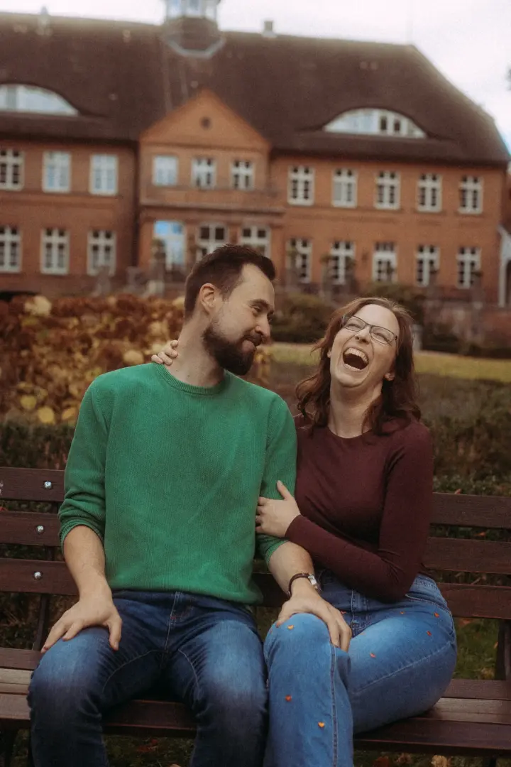 Ein Paar bei einem entspannten Fotoshooting: Ein Mann im grünen Pullover und eine lachende Frau mit Brille sitzen auf einer Parkbank vor einem historischen Backsteingebäude, festgehalten von einem Fotograf für Pärchen in Schwerin.