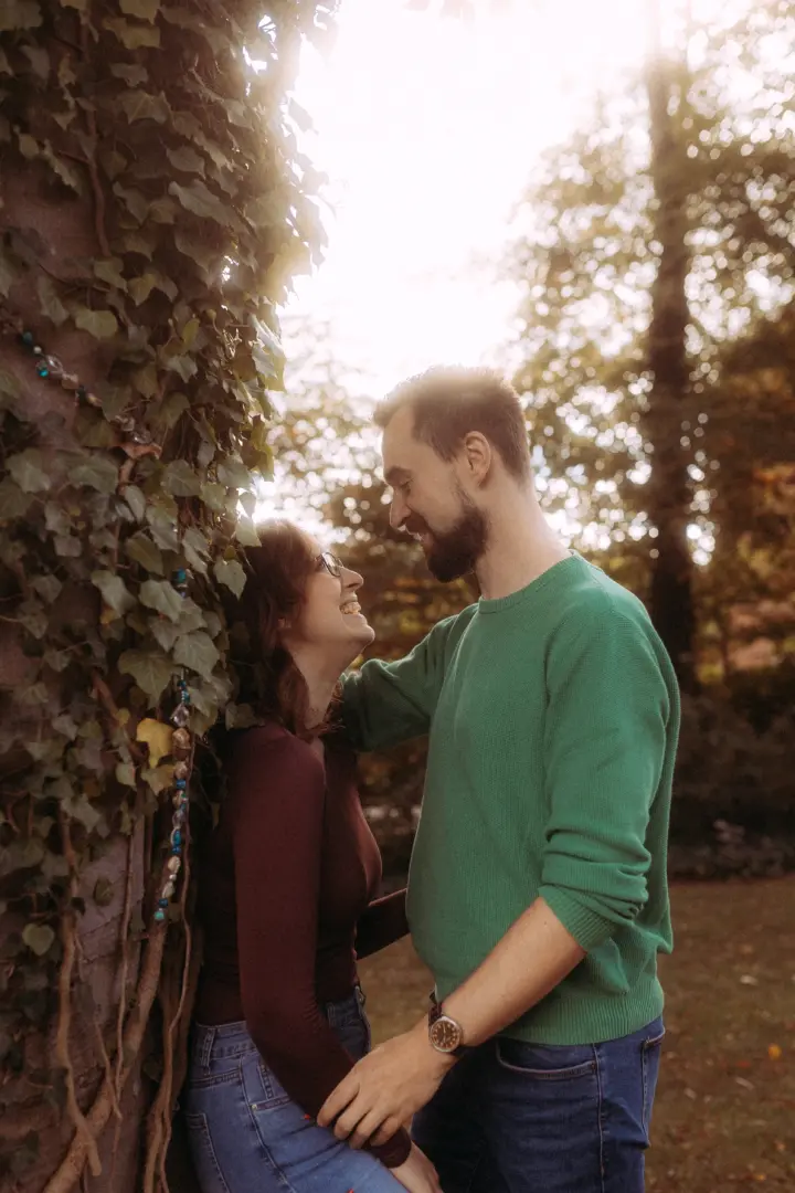 Ein glückliches Paar bei einem romantischen Fotoshooting im Herbstwald, bei dem sich beide liebevoll an einen mit Efeu bewachsenen Baum lehnen. Die warme Lichtstimmung unterstreicht die intime Atmosphäre dieser Pärchenfotos in Schwerin.