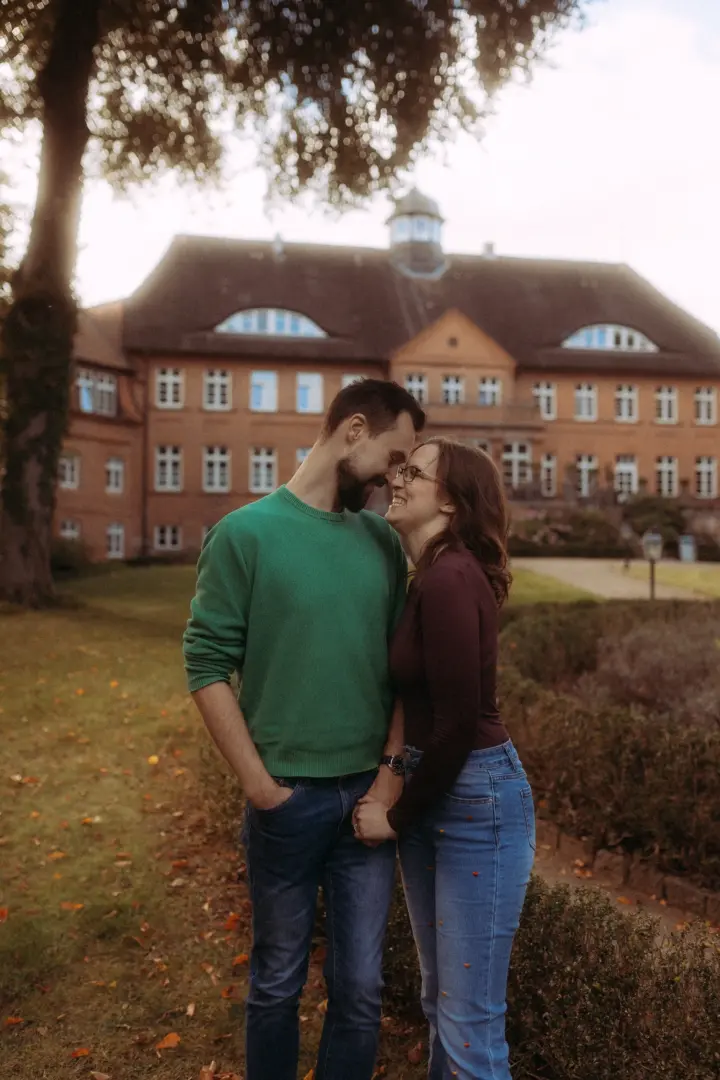 Ein verliebtes Paar steht sich bei einem romantischen Fotoshooting im Garten vor einem historischen Backsteingebäude gegenüber und hält Händchen. Die authentischen Pärchenfotos in Schwerin fangen die innige Verbindung zwischen den beiden in einer herbstlichen Atmosphäre ein.