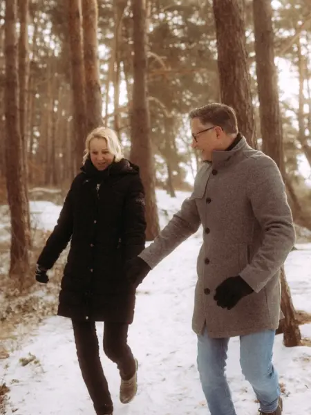 Ein glückliches Paar hält Händchen bei einem romantischen Winterspaziergang durch einen verschneiten Kiefernwald. Die beiden tragen warme Winterkleidung und genießen die gemeinsame Zeit in der Natur. Authentische Pärchenfotos Schwerin.