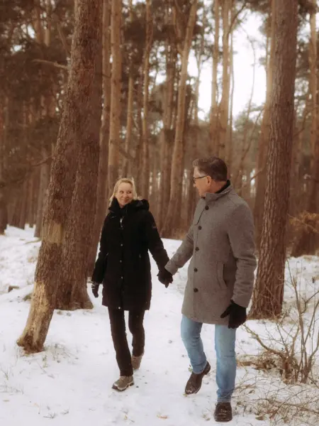 Ein glückliches Paar hält Händchen bei einem romantischen Winterspaziergang durch einen verschneiten Kiefernwald. Die beiden genießen die gemeinsame Zeit in der Natur, eingefangen von einem Fotograf für Pärchen in Schwerin.
