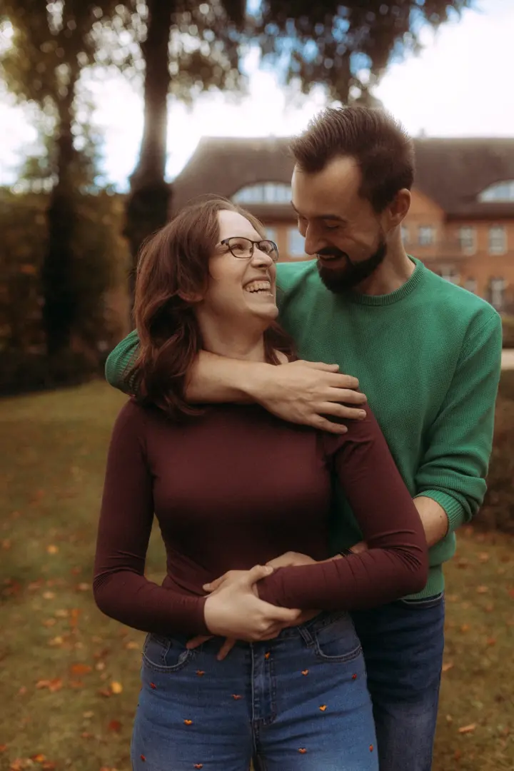Ein glückliches Paar bei einem Fotoshooting im Freien: Die Frau trägt ein weinrotes Langarmshirt und der Mann einen grünen Pullover, während sie sich liebevoll anlächeln. Diese natürlichen Pärchenfotos in Schwerin fangen einen intimen Moment vor einer herbstlichen Kulisse ein.
