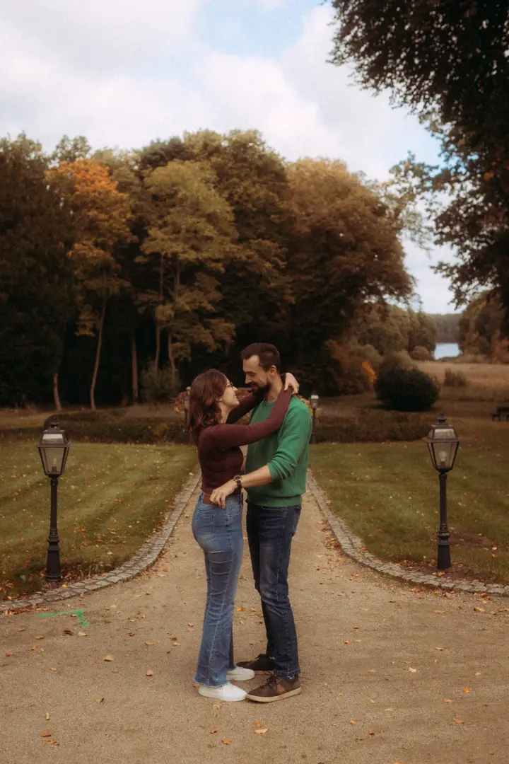 Ein verliebtes Paar in herbstlicher Kleidung steht sich auf einem von alten Laternen gesäumten Kiesweg in einem Park gegenüber. Die romantische Atmosphäre und die herbstliche Kulisse eignen sich perfekt für authentische Pärchenfotos Schwerin.
