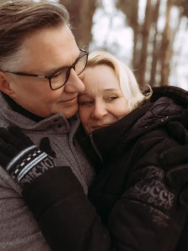 Ein liebevolles Paar in Winterkleidung umarmt sich eng im Freien. Der Mann trägt eine Brille und einen grauen Mantel, während die Frau in einer schwarzen Jacke an seine Schulter gelehnt ist. Ein stimmungsvolles Beispiel für authentische Pärchenfotos Schwerin in der Natur.