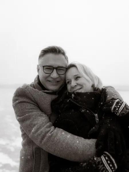 Ein glückliches Paar umarmt sich eng während eines Schneefalls im Winter. Der Mann trägt eine Brille und einen Mantel, die Frau eine dunkle Winterjacke und Handschuhe. Ein stimmungsvolles Bild für Pärchenfotos Schwerin, das die Verbundenheit der beiden in einer verschneiten Kulisse zeigt.