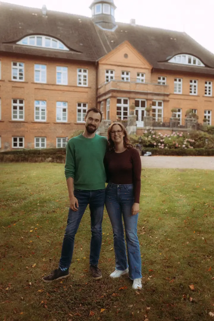 Ein glückliches Paar posiert für Pärchenfotos in Schwerin vor einem historischen Backsteingebäude im herbstlichen Garten. Der Mann trägt einen grünen Pullover, die Frau ein weinrotes Oberteil und Jeans. Die entspannte Atmosphäre unterstreicht die professionelle Arbeit als Fotograf für Pärchen.
