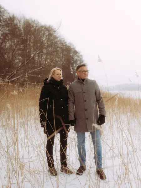 Ein Paar steht händchenhaltend in einer verschneiten Winterlandschaft mit hohem Schilf am Ufer eines zugefrorenen Sees. Die beiden tragen warme Winterkleidung und genießen die ruhige Atmosphäre. Professionelle Pärchenfotos Schwerin für unvergessliche Erinnerungen.