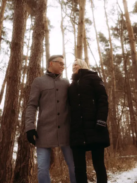 Ein glückliches Paar mittleren Alters genießt einen winterlichen Spaziergang im Wald. Der Mann trägt einen grauen Mantel und die Frau eine schwarze Steppjacke, während sie sich liebevoll ansehen. Authentische Pärchenfotos Schwerin in einer ruhigen, naturverbundenen Atmosphäre.
