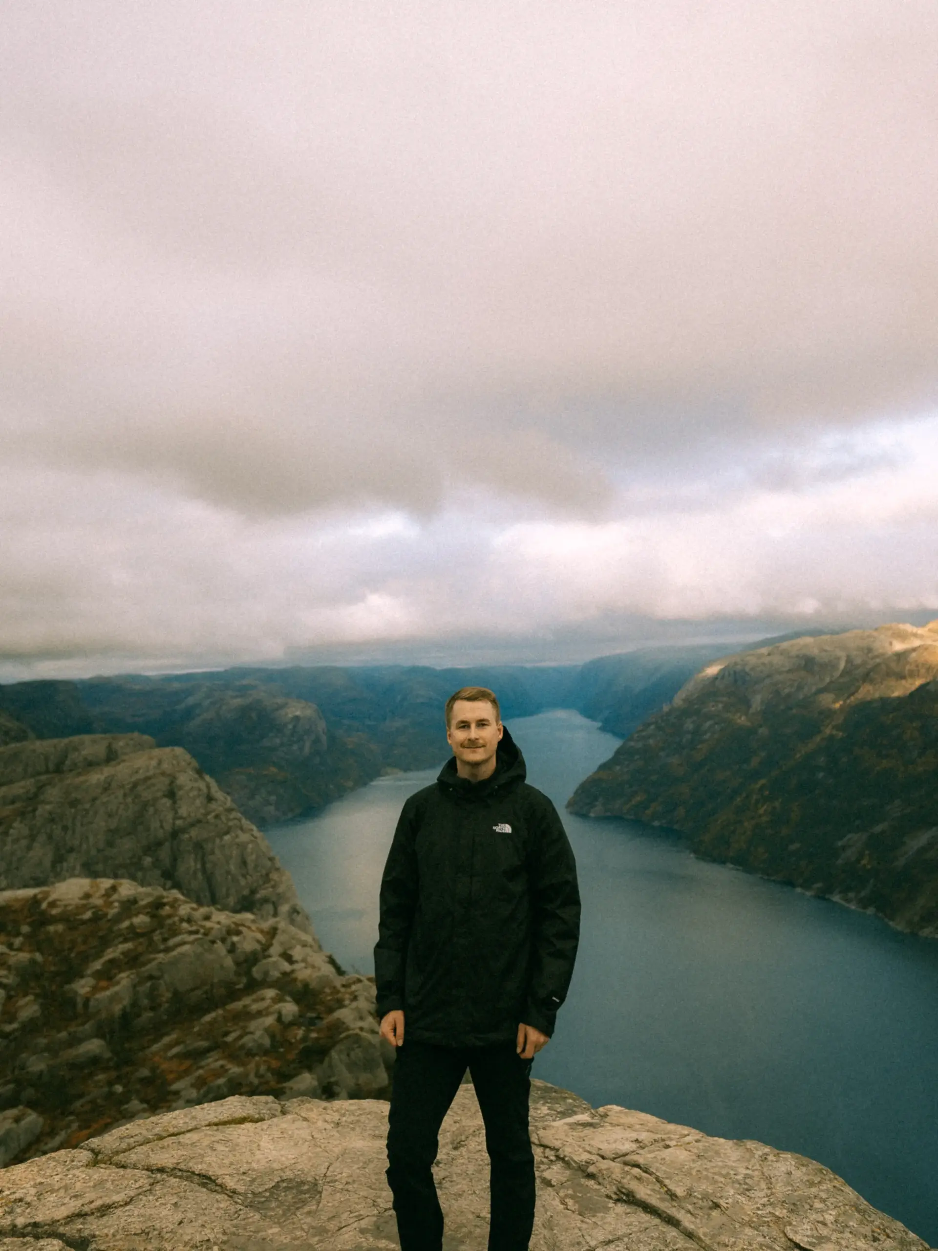 Ein junger Mann mit Schnurrbart in einer schwarzen Outdoor-Jacke steht auf einem felsigen Gipfelplateau vor dem Lysefjord in Norwegen. Im Hintergrund erstreckt sich das tiefblaue Wasser des Fjords zwischen steilen, herbstlich gefärbten Felswänden unter einem bewölkten Himmel. Ein junger Mann mit Schnurrbart in einer schwarzen Outdoor-Jacke steht auf einem felsigen Gipfelplateau vor dem Lysefjord in Norwegen. Im Hintergrund erstreckt sich das tiefblaue Wasser des Fjords zwischen steilen, herbstlich gefärbten Felswänden unter einem bewölkten Himmel.