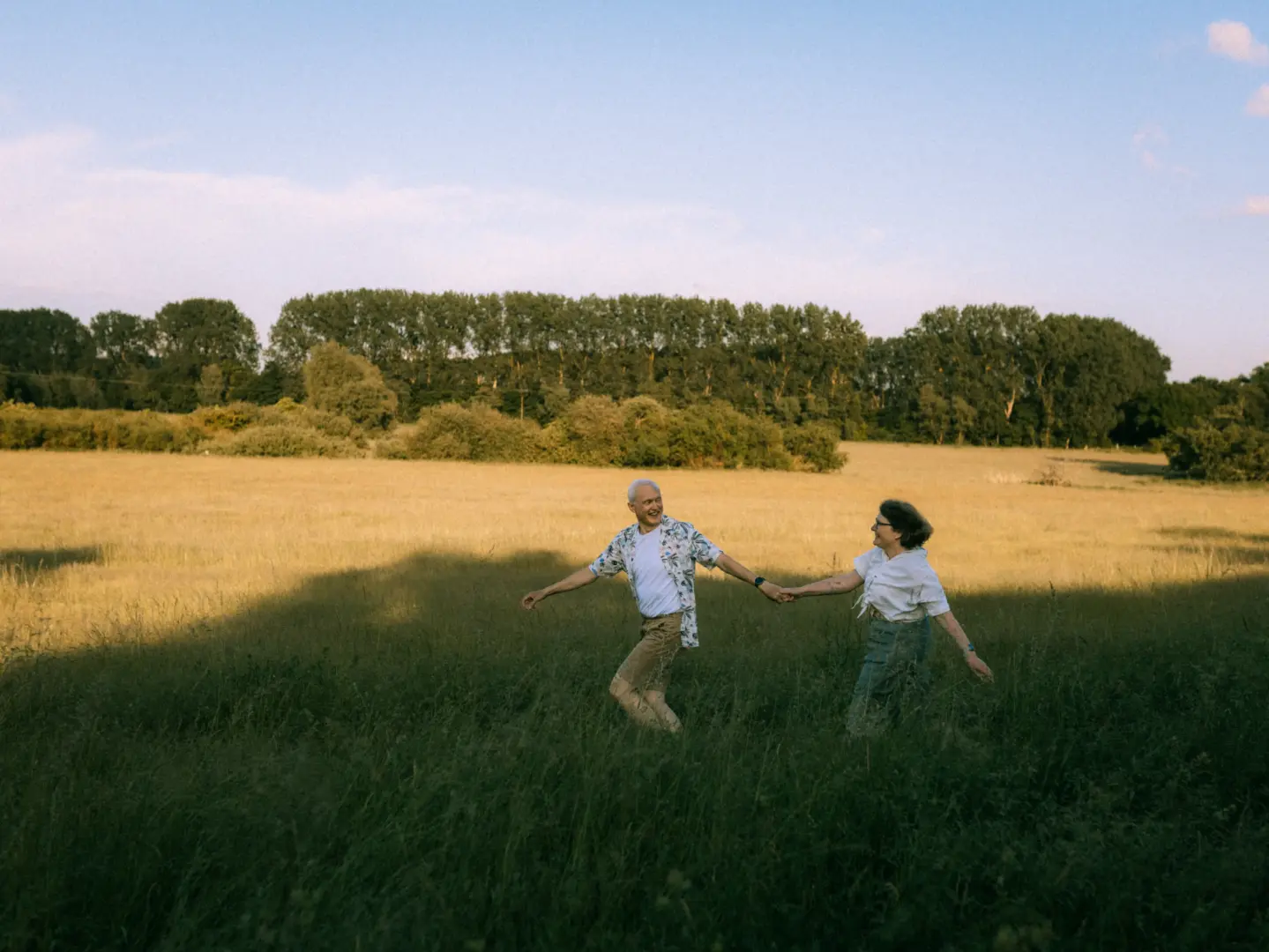 Ein älteres Paar hält händchenhaltend und lachend inne beim Spaziergang durch eine sonnendurchflutete Sommerwiese. Die beiden wirken glücklich und entspannt in dieser natürlichen Umgebung. Professionelle Pärchenfotos Schwerin, die echte Emotionen einfangen.