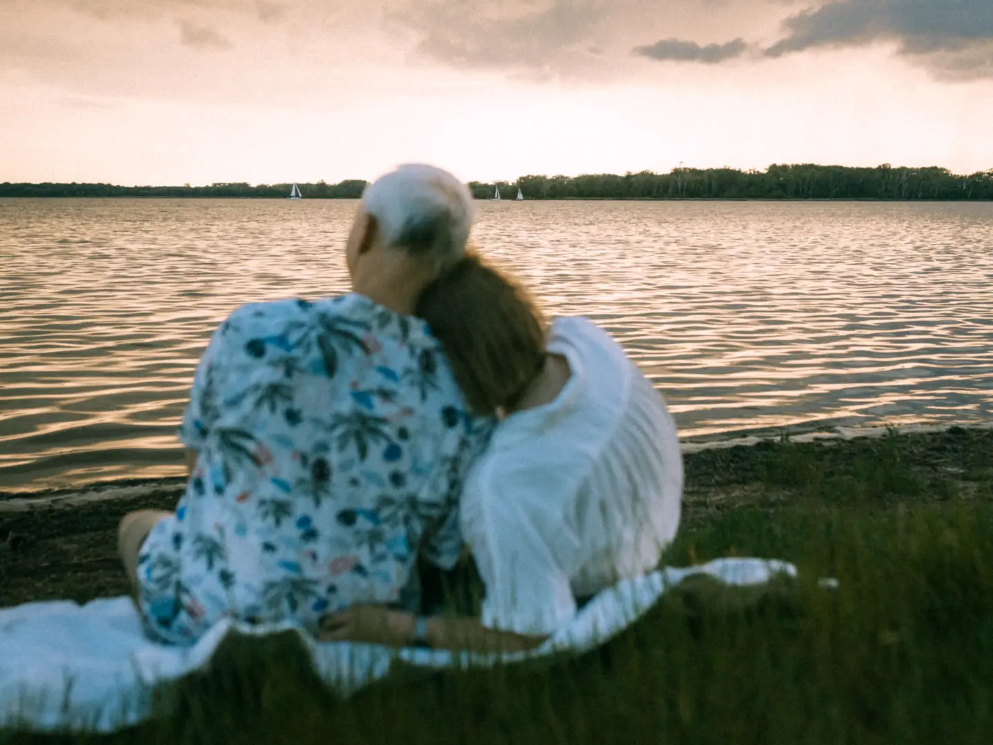 Ein Paar sitzt eng umschlungen auf einer Decke am Ufer eines Sees und genießt den Blick auf das Wasser bei Sonnenuntergang. Im Hintergrund segeln kleine Boote in der Ferne. Romantische Pärchenfotos Schwerin, eingefangen in einer stimmungsvollen Abendatmosphäre.