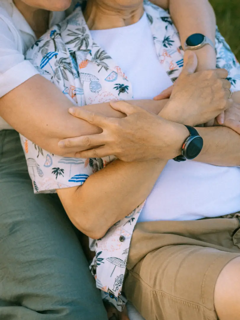 Ein Paar in sommerlicher Kleidung bei einer innigen Umarmung: Die Person im Vordergrund trägt ein weißes T-Shirt unter einem kurzärmeligen Hemd mit Palmen- und Sonnenschirm-Muster sowie eine beigefarbene Shorts. Emotionale Pärchenfotos Schwerin in einer entspannten, sommerlichen Atmosphäre.