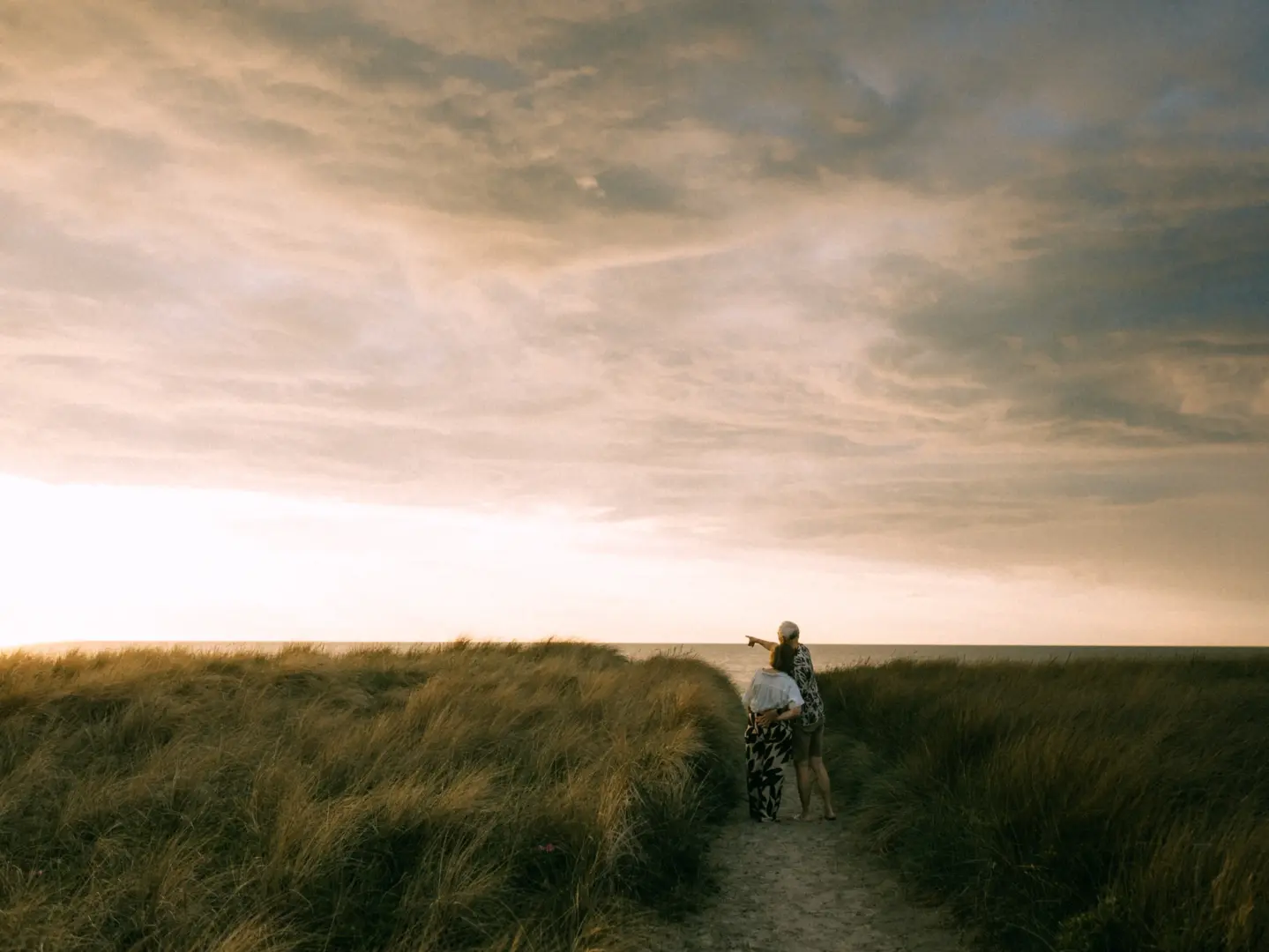 Ein Paar steht Arm in Arm auf einem Sandweg in den Dünen und blickt gemeinsam auf das weite Meer bei Sonnenuntergang. Stimmungsvolle Pärchenfotos Schwerin in einer idyllischen Küstenlandschaft mit goldenem Licht und bewölktem Himmel.