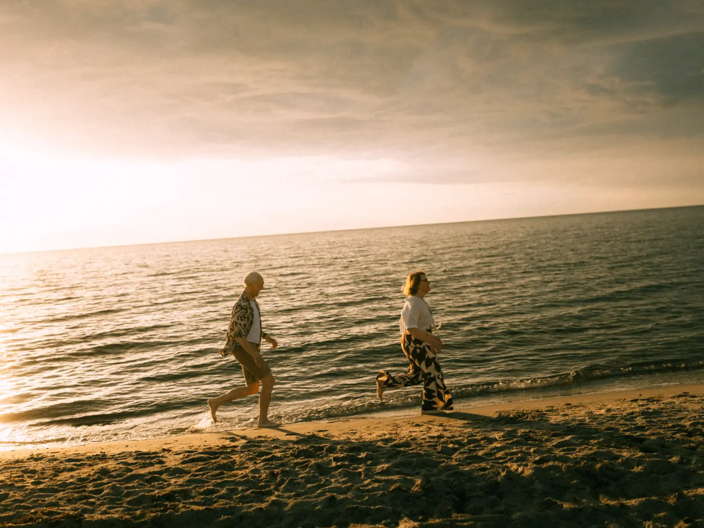 Ein älteres Paar läuft barfuß und glücklich am sandigen Strand entlang, während die warme Abendsonne das Meer in goldenes Licht taucht. Die beiden genießen einen entspannten Moment am Wasser. Lovestory Fotograf in Schwerin und MV.