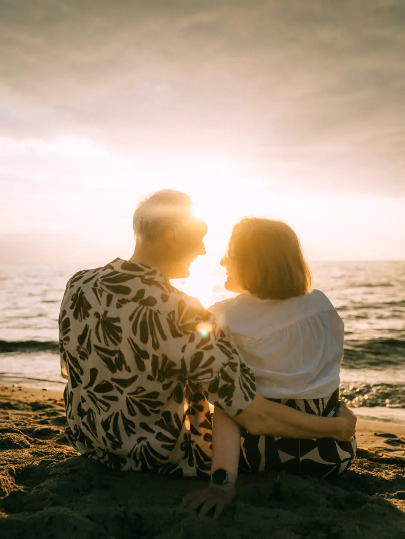 Ein Paar sitzt eng umschlungen im Sand am Strand und blickt sich bei einem stimmungsvollen Sonnenuntergang am Meer liebevoll in die Augen. Diese romantischen Pärchenfotos Schwerin fangen die warme Abendstimmung und ihre tiefe Verbundenheit perfekt ein.