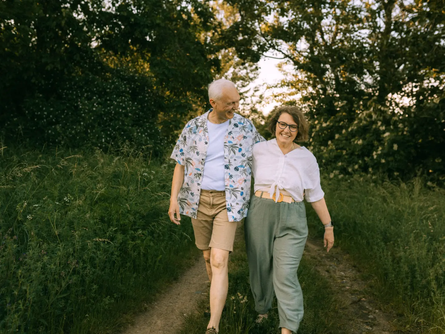 Ein glückliches älteres Paar spaziert bei warmem Abendlicht Arm in Arm auf einem Feldweg durch die Natur. Der Mann trägt ein buntes Hawaiihemd, die Frau eine helle Bluse und eine grüne Hose. Authentische Pärchenfotos Schwerin, die eine liebevolle Verbindung in der Natur zeigen.