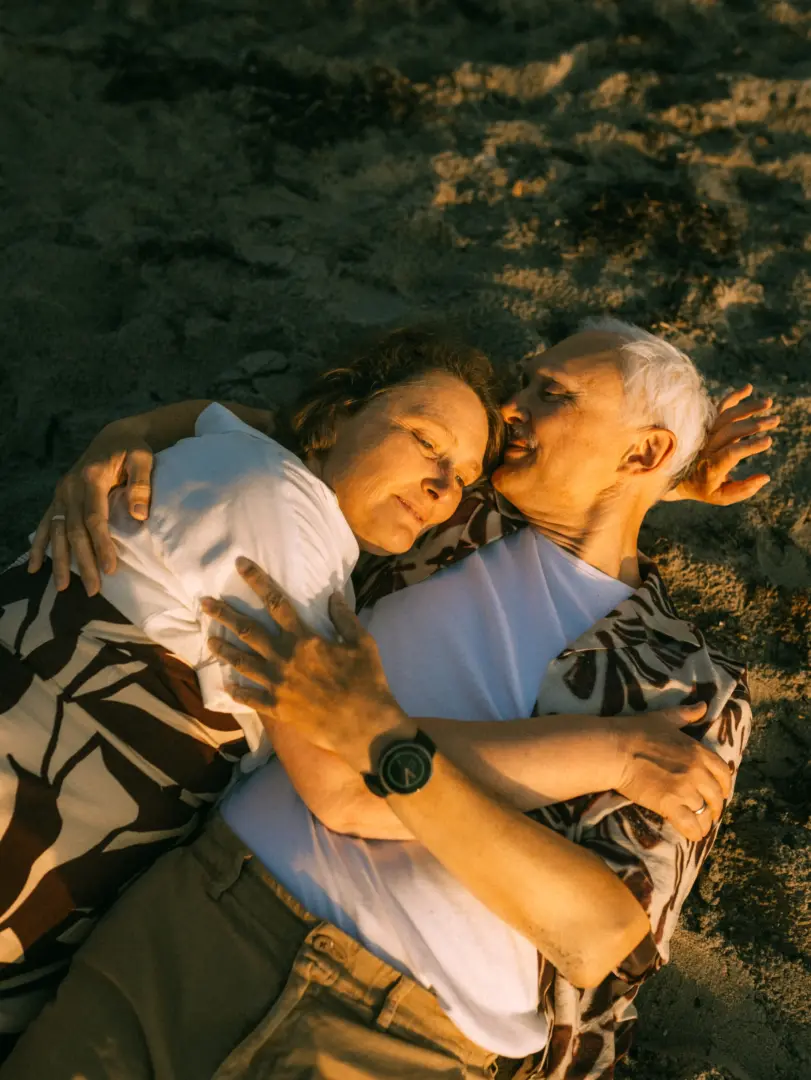 Ein älteres Paar liegt sich liebevoll im Arm auf dem Sand am Strand, während die warme Abendsonne ihre Gesichter in goldenes Licht taucht. Die beiden wirken glücklich und entspannt bei diesen natürlichen Pärchenfotos Schwerin.
