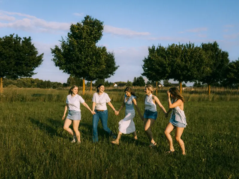 Fünf junge Frauen lachen fröhlich, während sie sich an den Händen haltend in einer Reihe über eine sonnige Wiese laufen. Die Szene strahlt Lebensfreude aus, perfekt eingefangen für natürliche Freundschaftsfotos oder als Inspiration für ein Fotoshooting in Schwerin.