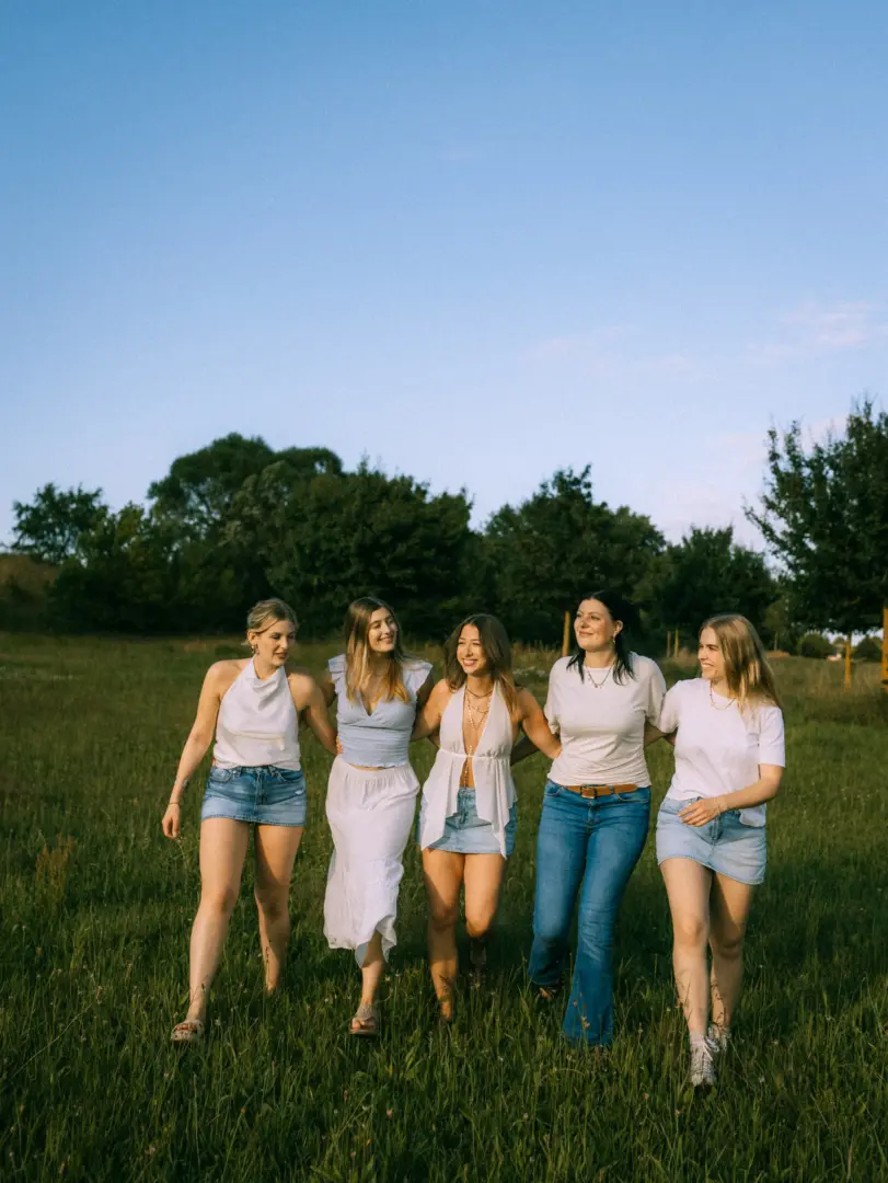 Fünf junge Frauen gehen lachend und Arm in Arm über eine sonnige Wiese. Sie tragen sommerliche Outfits in Weiß- und Blautönen und genießen die entspannte Atmosphäre im Freien. Das warme Licht des späten Nachmittags unterstreicht die freundschaftliche Stimmung in dieser natürlichen Kulisse.