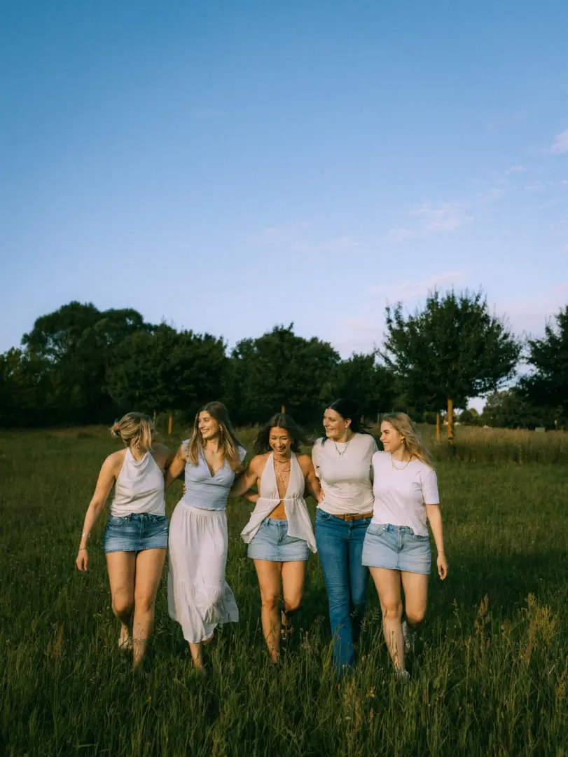Fünf junge Frauen lachen gemeinsam und laufen Arm in Arm durch eine sonnige Wiese. Sie tragen sommerliche Outfits in Weiß und Blau, während sie im warmen Abendlicht vor einer Kulisse aus Bäumen spazieren. Ein stimmungsvoller Moment für authentische Freundschaftsfotos.