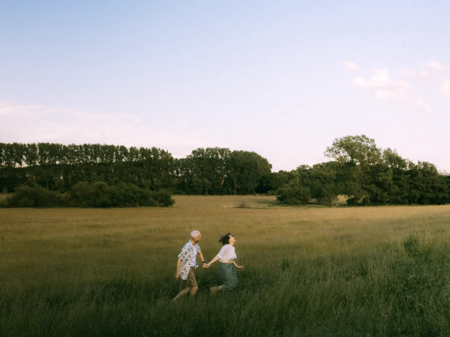 Ein glückliches Paar rennt lachend und Händchen haltend durch eine weite, goldene Wiese bei Sonnenuntergang. Die beiden genießen den Moment in der Natur, während im Hintergrund ein Waldrand die Szenerie abrundet. Authentische Pärchenfotos Schwerin für unvergessliche Erinnerungen.