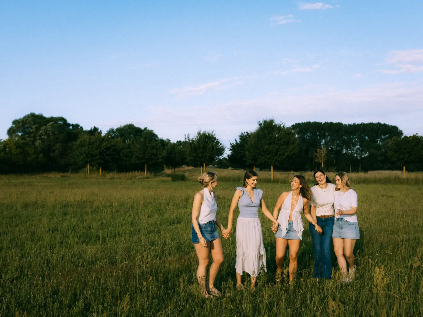 Vier junge Frauen halten sich lachend an den Händen und spazieren gemeinsam durch eine sonnendurchflutete Wiese bei Sonnenuntergang. Sie tragen sommerliche Outfits in Weiß und Blau, während im Hintergrund eine Baumreihe unter einem klaren Abendhimmel zu sehen ist.