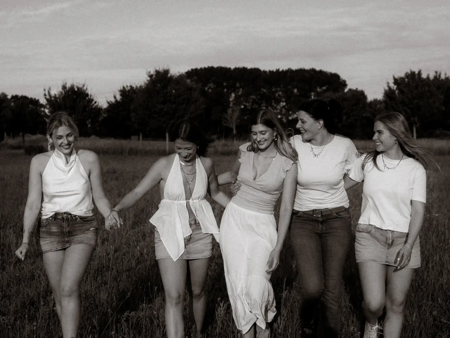 Fünf junge Frauen lachen gemeinsam, während sie Hand in Hand durch ein sonniges Feld spazieren. Die schwarz-weiße Aufnahme fängt eine fröhliche Stimmung ein, als sie sich in sommerlicher Kleidung entspannt und freundschaftlich durch die Natur bewegen.