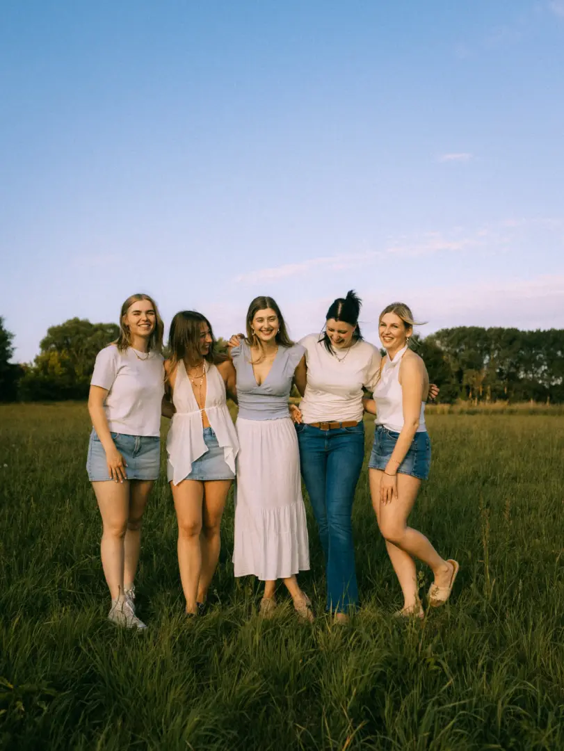 Fünf junge Frauen stehen eng umschlungen und lächelnd auf einer sonnendurchfluteten Wiese bei Sonnenuntergang. Sie tragen sommerliche Outfits in Weiß und Blau. Das freundschaftliche Gruppenbild strahlt Lebensfreude aus und eignet sich ideal als Inspiration für ein Fotoshooting.