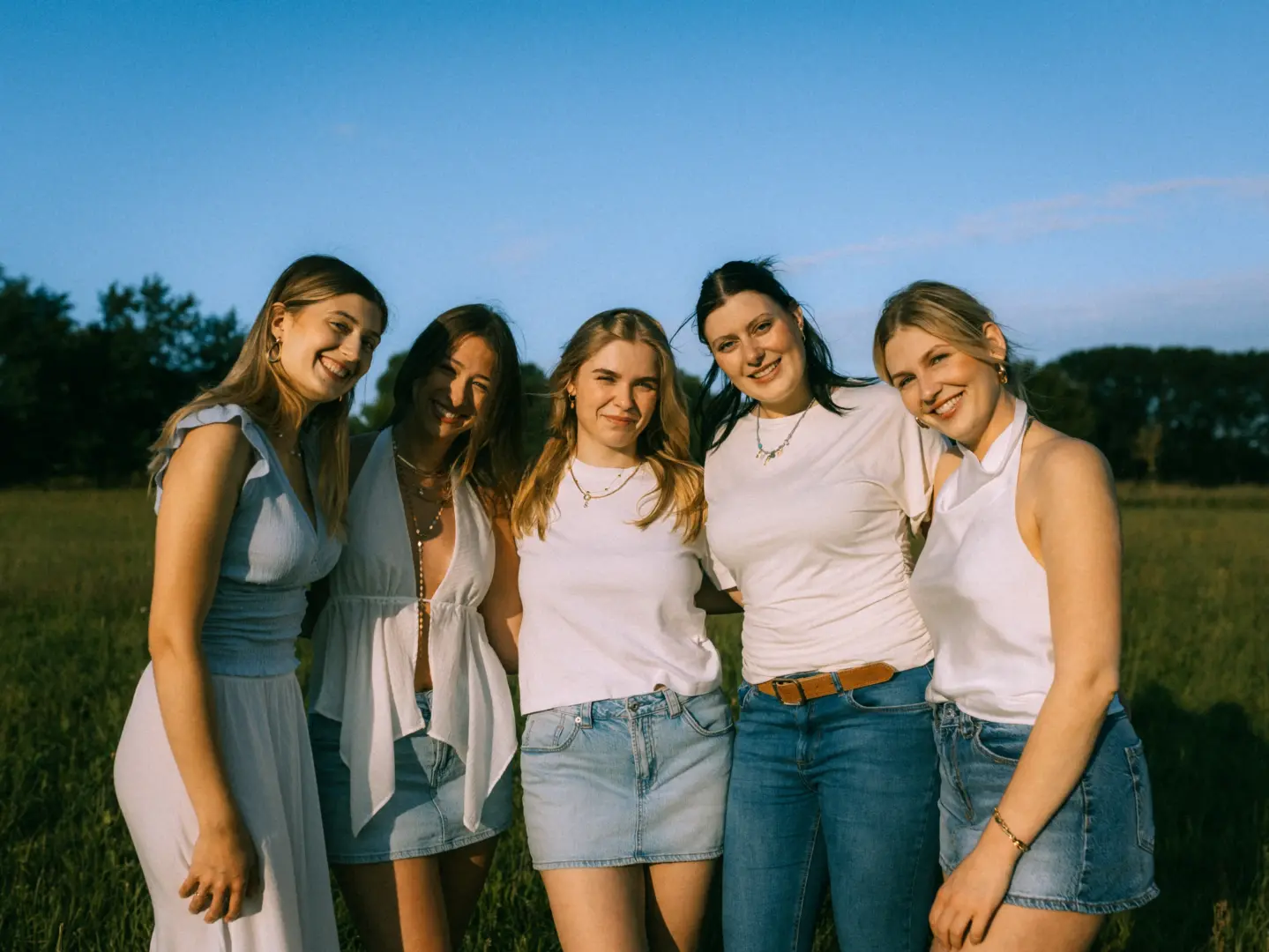 Fünf junge Frauen stehen eng beieinander auf einer Wiese im warmen Licht der Abendsonne und lächeln in die Kamera. Sie tragen sommerliche, helle Outfits in Weiß und Blau, während im Hintergrund Bäume unter einem klaren blauen Himmel zu sehen sind. Ein stimmungsvolles Gruppenfoto.
