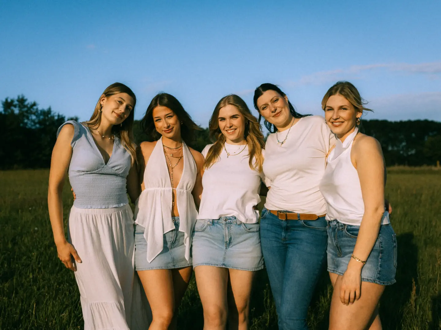 Fünf junge Frauen stehen eng beieinander auf einer sonnigen Wiese bei goldenem Abendlicht. Sie tragen sommerliche Outfits in Weiß und Blau, lächeln freundlich in die Kamera und strahlen eine herzliche Freundschaft aus. Im Hintergrund erstreckt sich eine weite, natürliche Landschaft.