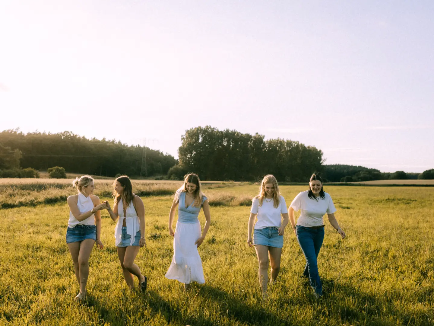Fünf junge Frauen spazieren lachend und gut gelaunt durch eine sonnige Sommerwiese. Sie tragen legere, helle Kleidung wie Jeansröcke und weiße Tops, während das warme Abendlicht eine harmonische Atmosphäre schafft. Dieses Bild entstand bei einem Shooting als Fotograf für Pärchen in Schwerin.
