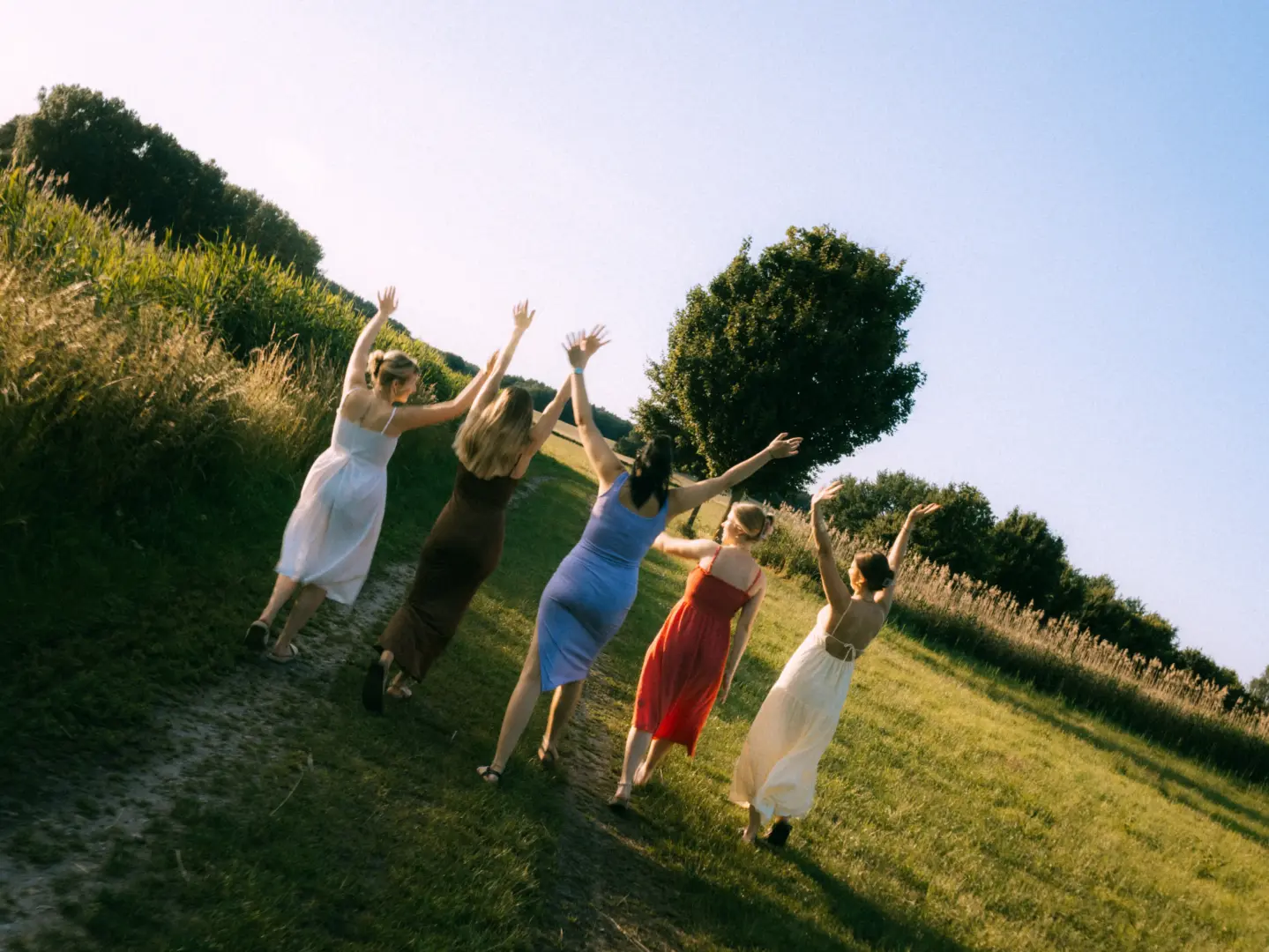 Fünf junge Frauen in eleganten Sommerkleidern laufen bei Sonnenuntergang fröhlich einen Feldweg entlang und strecken ihre Arme in den Himmel. Die Szene strahlt Freiheit und Lebensfreude aus. Hochzeitsfotograf in Schwerin und MV.
