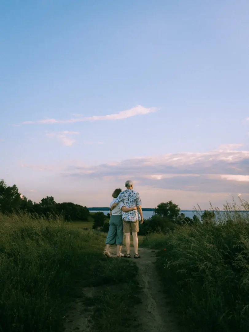 Ein älteres Paar steht Arm in Arm auf einem Feldweg und blickt gemeinsam auf das ruhige Wasser am Horizont. Die Szene in der Abenddämmerung fängt einen romantischen Moment ein, ideal für Pärchenfotos Schwerin in einer idyllischen Naturkulisse.
