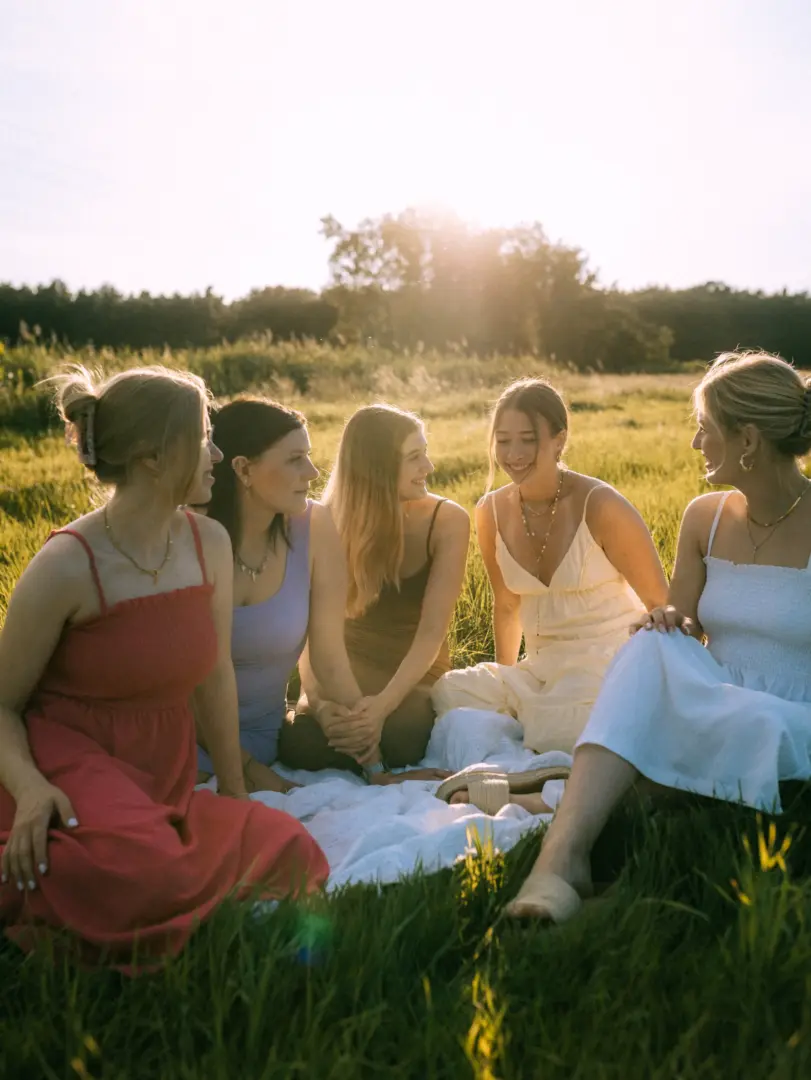 Fünf junge Frauen sitzen bei goldenem Sonnenuntergang auf einer Decke in einer Wiese und unterhalten sich fröhlich. Die Szene strahlt eine warme, sommerliche Atmosphäre aus, während die Frauen in hellen, pastellfarbenen Sommerkleidern entspannt die gemeinsame Zeit in der Natur genießen.