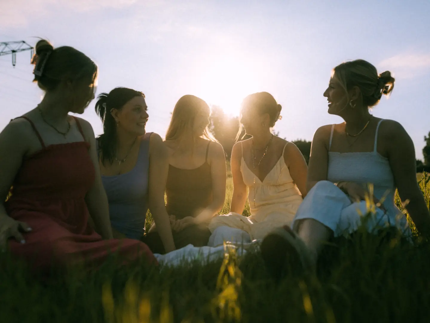 Fünf junge Frauen sitzen bei sommerlichem Sonnenuntergang gemeinsam auf einer Wiese und unterhalten sich fröhlich. Die Szene ist in warmes Gegenlicht getaucht, was eine freundschaftliche und entspannte Stimmung erzeugt. Professionelle Pärchenfotos Schwerin für besondere Momente.