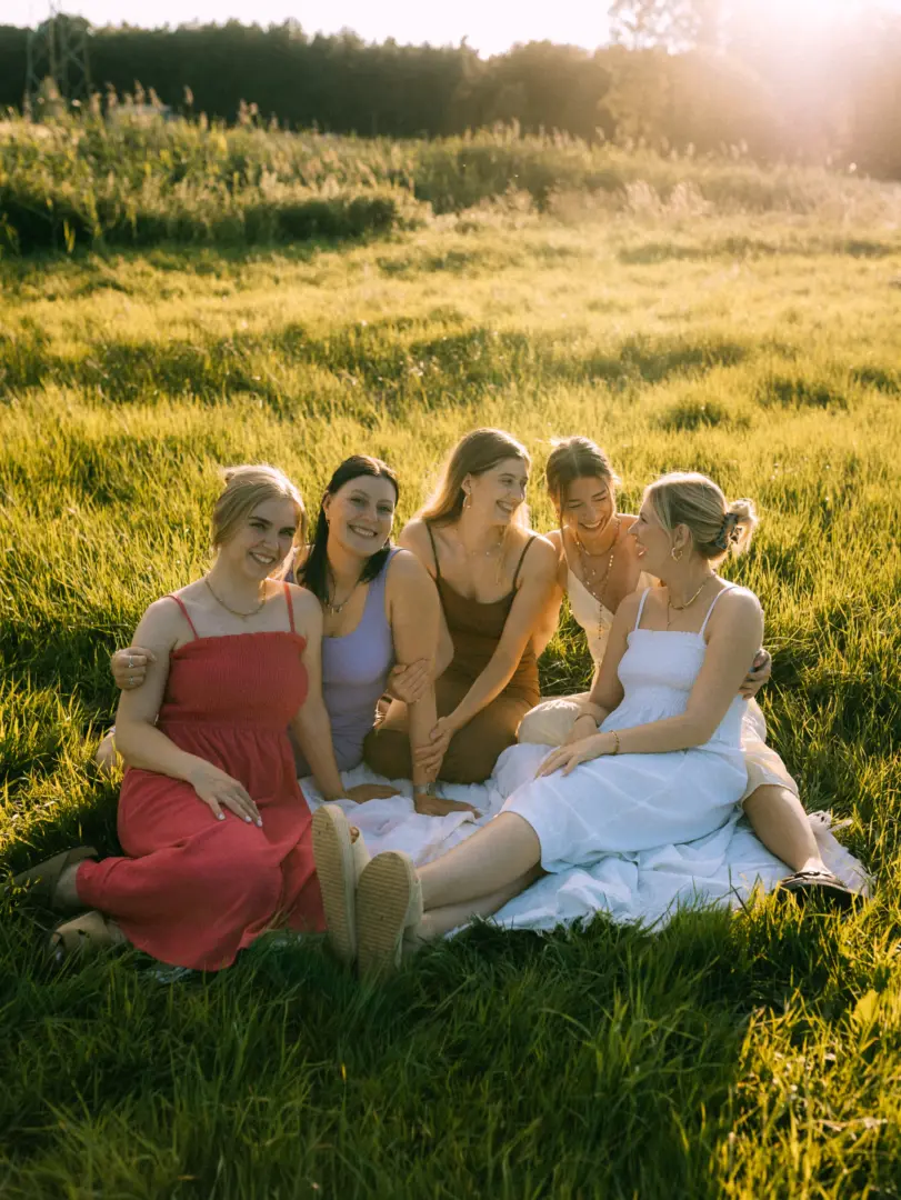 Fünf junge Frauen sitzen gemeinsam auf einer Picknickdecke in einer sonnendurchfluteten Wiese. Sie tragen sommerliche Kleider in verschiedenen Farben, lachen herzlich und genießen die warme Abendstimmung. Ein authentischer Moment, ideal für ein Fotoshooting oder Pärchenfotos Schwerin.