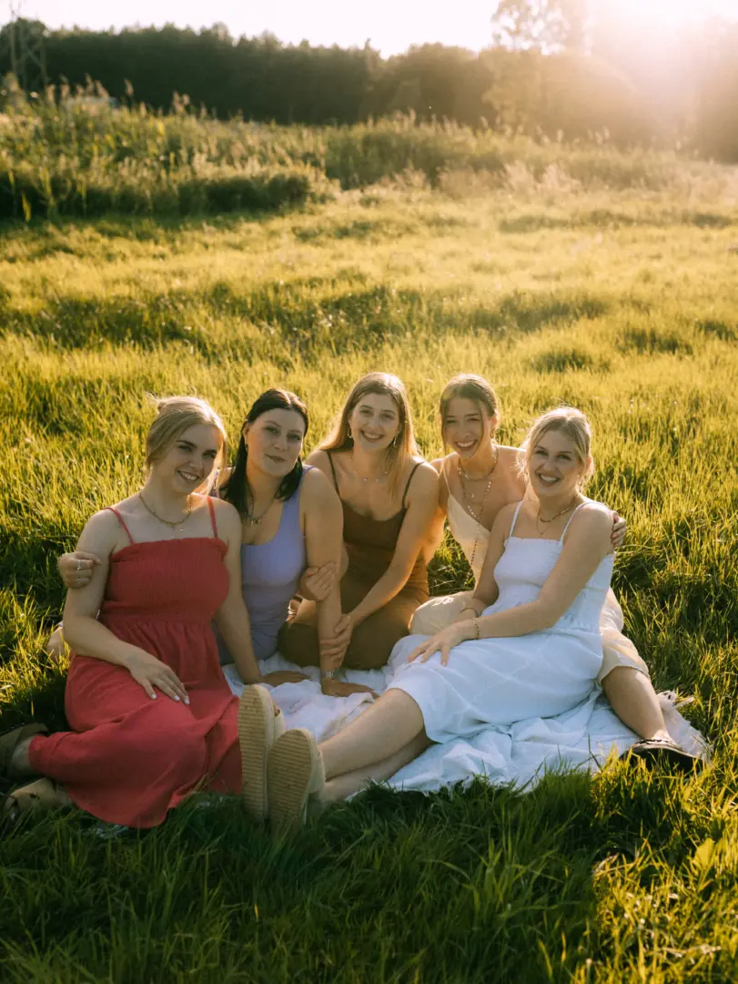 Fünf junge Frauen sitzen gemeinsam auf einer weißen Decke in einer sonnendurchfluteten Wiese. Sie lächeln in die Kamera, tragen sommerliche Kleider in verschiedenen Farben und genießen die warme Abendstimmung. Ein herzliches Bild für Familienfotos in Schwerin.