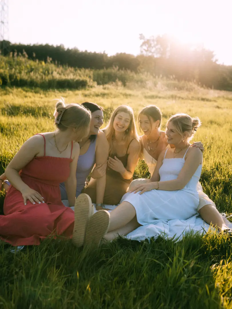 Fünf junge Frauen lachen gemeinsam bei einem Picknick auf einer sonnigen Wiese während des Golden Hour-Lichts. Sie tragen sommerliche Kleider in Pastellfarben und Weiß. Diese Aufnahme entstand bei einem Fotoshooting, wie es auch ein Fotograf für Pärchen in Schwerin anbietet.