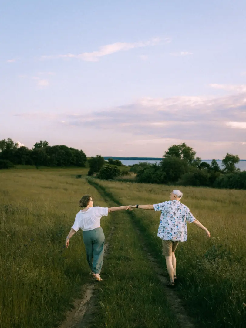 Ein Paar hält sich an den Händen und läuft fröhlich einen Feldweg entlang in Richtung eines Sees. Die Szene fängt eine sommerliche Stimmung ein, ideal für authentische Pärchenfotos Schwerin, bei denen die Verbundenheit in der Natur im Fokus steht.