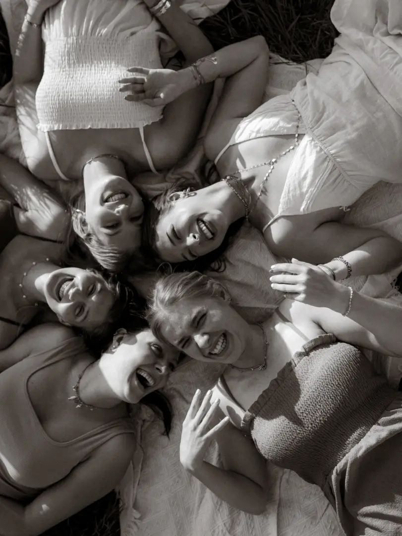 Vier junge Frauen liegen in einer sternförmigen Anordnung auf einer hellen Decke im Freien. Sie lachen herzlich in die Kamera, während sie die Sonne genießen. Die schwarz-weiße Aufnahme fängt einen fröhlichen Moment unter Freunden ein, ideal als Inspiration für authentische Pärchenfotos Schwerin.