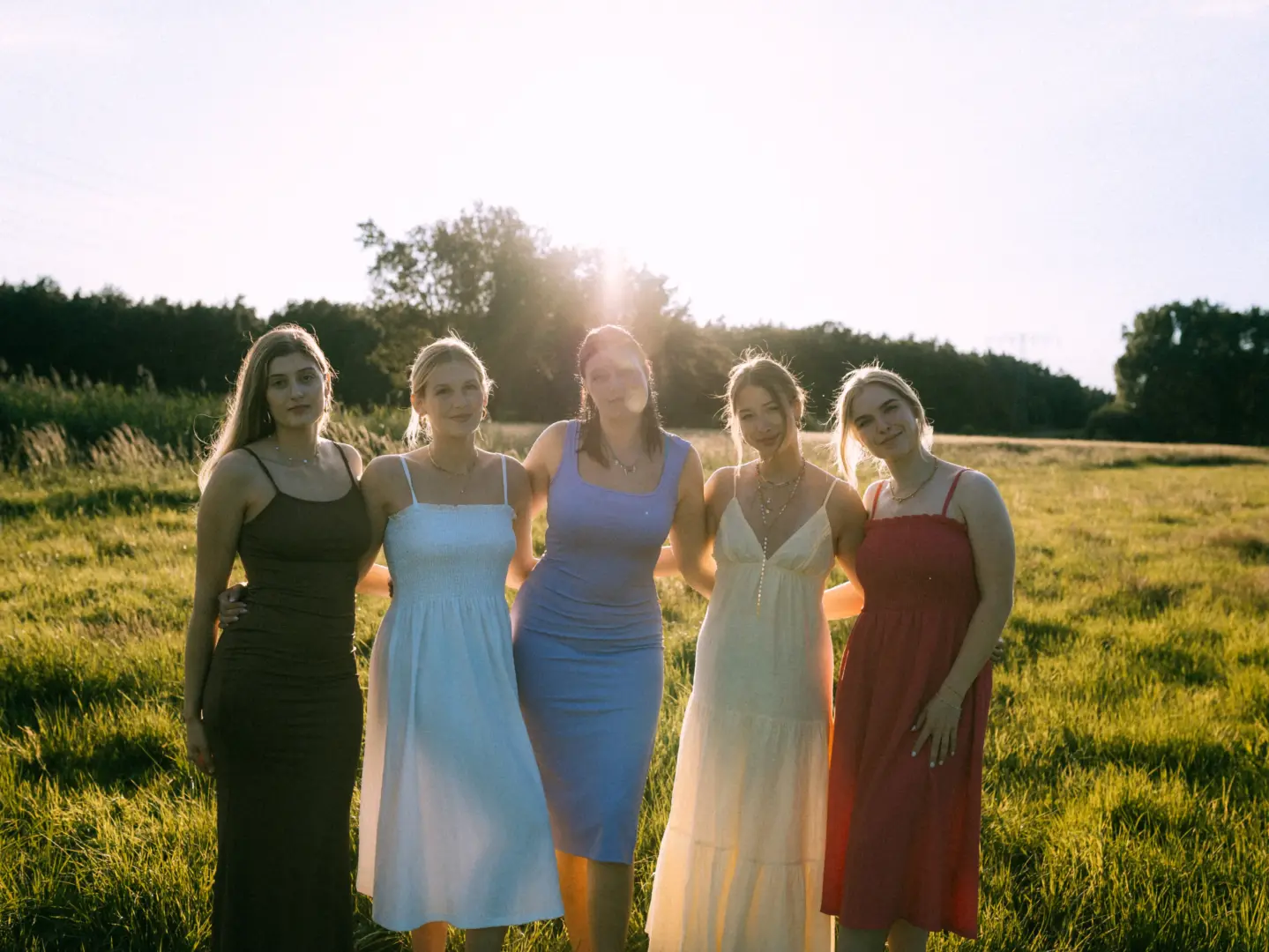 Fünf junge Frauen in sommerlichen Kleidern stehen eng beieinander auf einer sonnigen Wiese während des goldenen Lichts. Die Szene strahlt Freundschaft und Lebensfreude aus, perfekt eingefangen von einem Fotografen für Pärchen und Gruppen in Schwerin.
