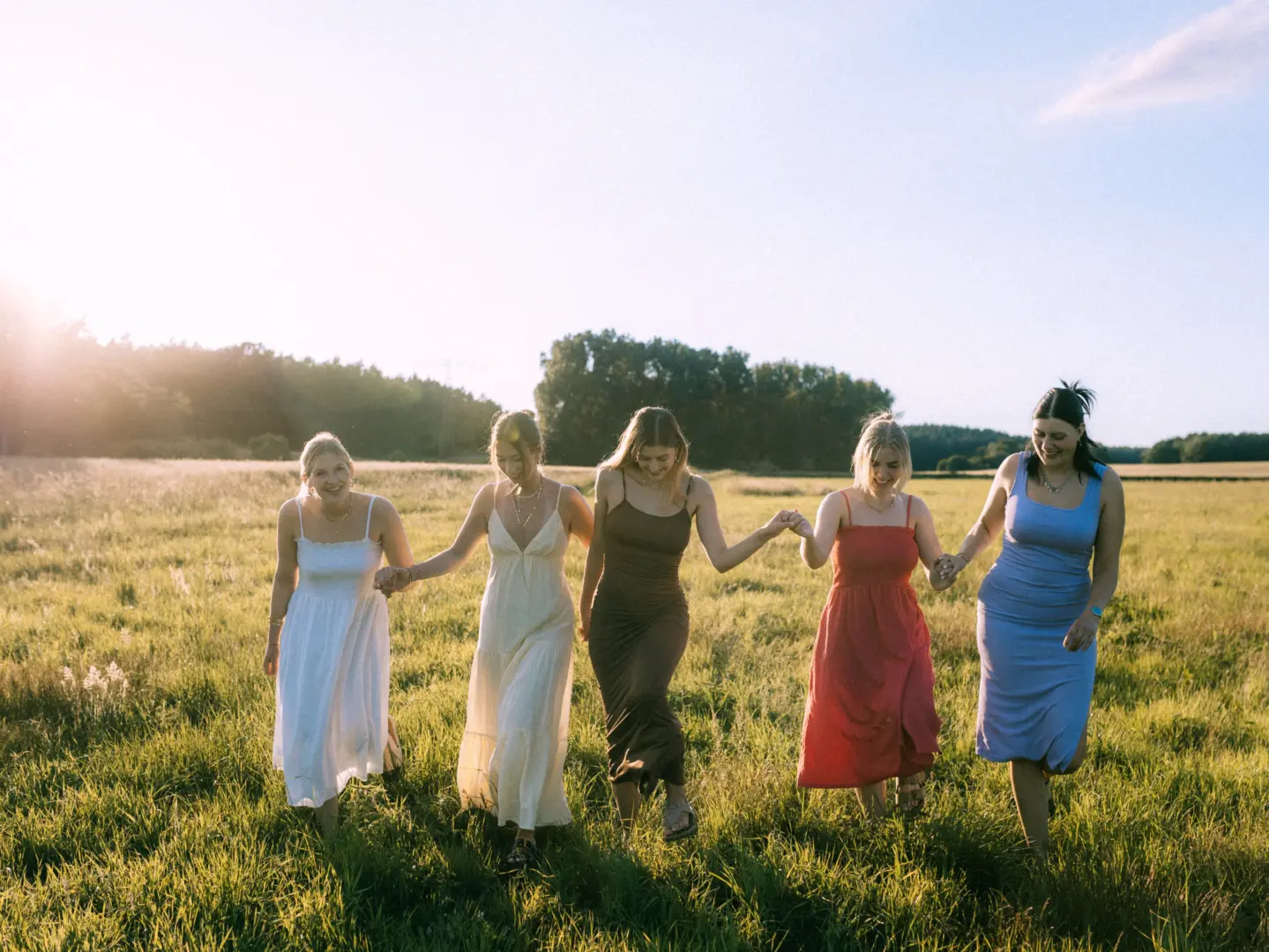 Fünf junge Frauen in sommerlichen Kleidern gehen Hand in Hand lachend über eine sonnendurchflutete Wiese bei Sonnenuntergang. Die Szene strahlt Lebensfreude und Verbundenheit aus. Ein herzlicher Moment, festgehalten von Ihrem Fotograf für Pärchen in Schwerin.